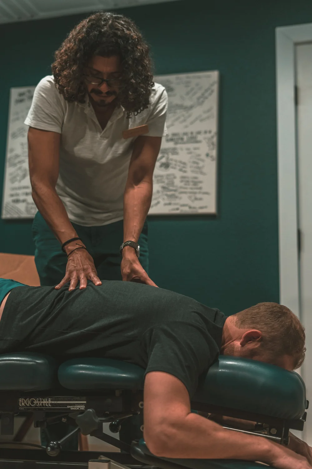 Nurse Doza standing beside a padded treatment table in a dark green treatment room, both hands resting on the upper back of a person lying face down.