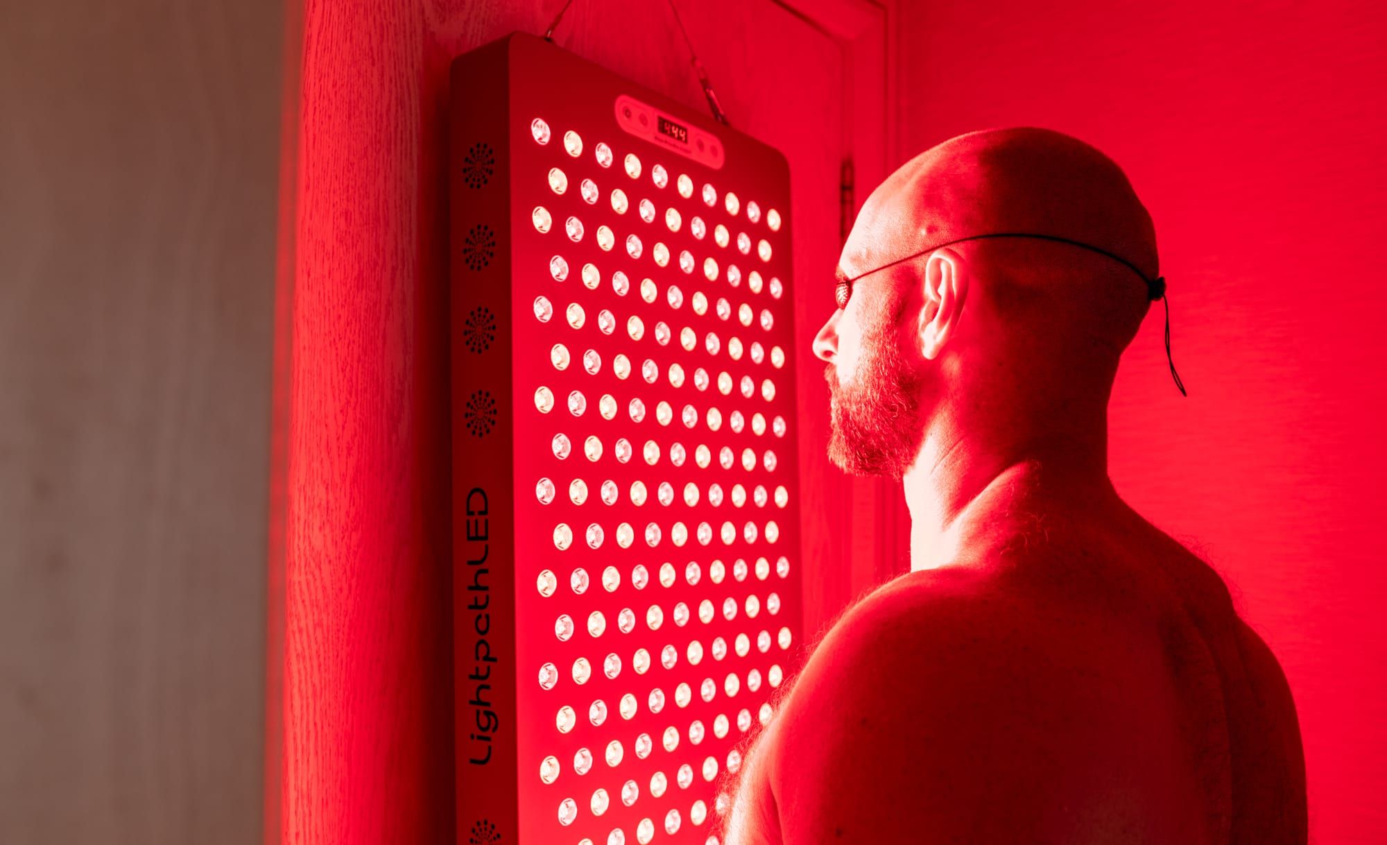 A wall-mounted red light panel with rows of illuminated LEDs, and a person standing beside it in profile wearing protective eyewear.