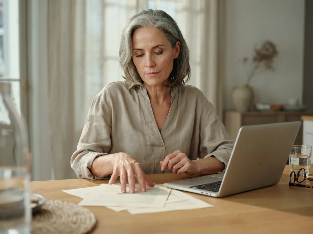 A woman reviewing a printed document at a kitchen table beside a laptop.